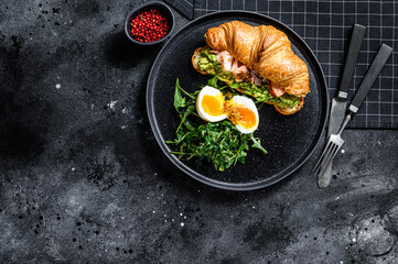 Croissant with hot smoked salmon, avacado, arugula and egg. Black background. Top view. Space for text