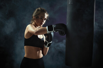 sporty brunette woman in boxing gloves and sportswear kicking bag on dark background with smoke.