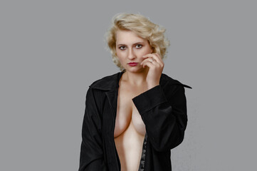 Blonde girl with a naked breast in a black cloak looks at the camera seductively on a gray background
