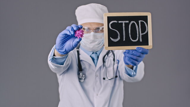 Male lab technician in medical uniform, protective mask and glasses shows covid-19 virus cell and stop sign in research laboratory on white background. Close-up view focused on coronavirus cell