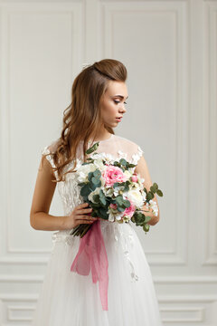 portrait of beautiful tender bride with wedding makeup and long curly hair holding flower bouquet in studio interior