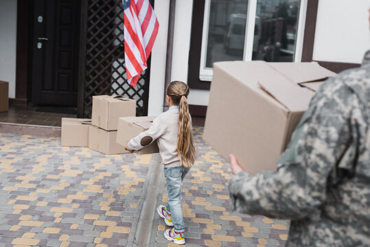 Back view of girl carrying cardboard box near house with blurred father on foreground