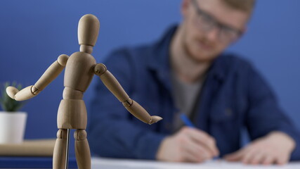 Obraz premium Designer artist paints a wooden gestalt figure. The work of the artist or designer. The creative process. Creative workspace in blue tones. A man with glasses is copying on paper a wooden man. Blur