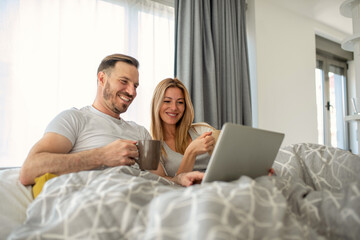 Fototapeta premium Young couple sitting in bed in the morning, reading news on laptop and drinking coffee together