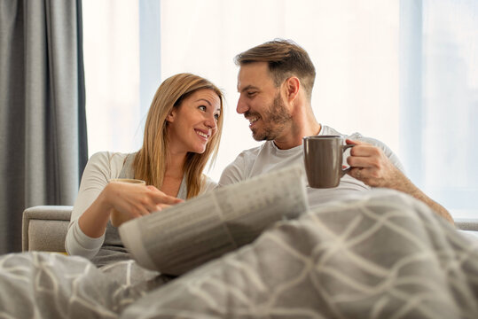 Beautiful young couple in love, sitting in bed in the morning, reading newspapers and drinking coffee together