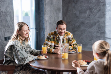 Happy couple having breakfast, laughing, while sitting at table in kitchen with blurred girl on foreground