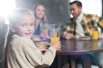Happy girl looking at camera, while holding glass of juice and sitting at table with blurred woman and man on background