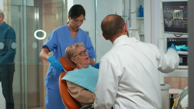 Nurse putting dental bib to old woman during stomatological examination. Doctor and nurse working together in modern orthodontic clinic showing radiography of teeth on monitor pointing on digital