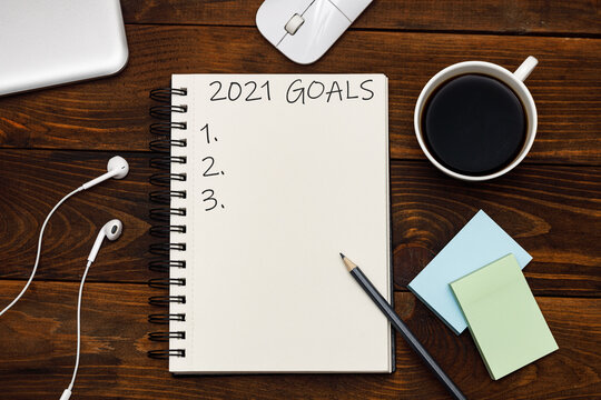2021 goals text on notepad, chek list. Top view of headphones, mouse, coffee and notebook on wooden background. Flat lay.