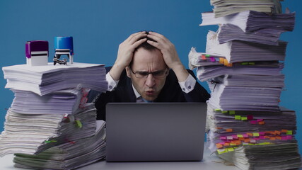 Concentrated office worker businessman working on laptop among piles of paperwork and stamps on stack of documents overloaded with work. Grabbing his head because of an mistake, deadline concept