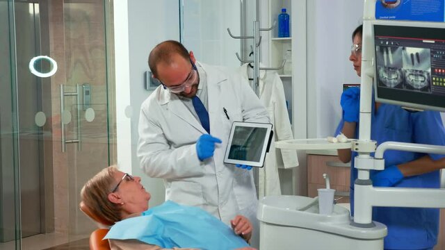 Dentist in dental office talking with woman patient and preparing for treatment, examining x-ray image on tablet. Doctor showing to old woman dental radiography modern gadget in stomatological clinic.