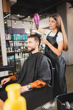 young hairdresser in apron drying hair of smiling man, blurred foreground