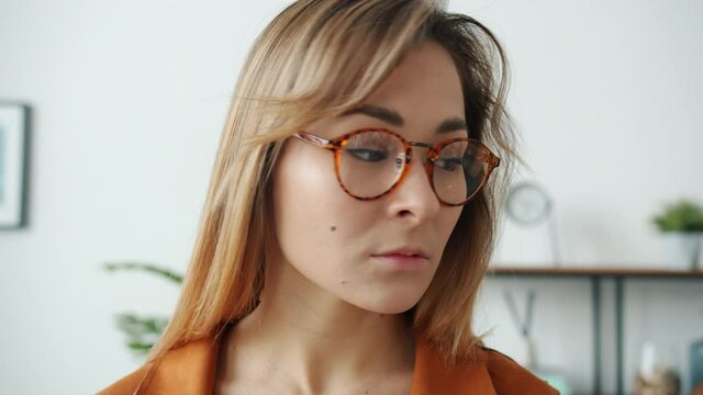 Portrait of serious young blond lady wearing trendy glasses looking at camera standing alone at home. Human emotions and modern youth concept.