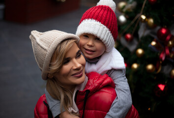 Happy family, young pretty woman with child spend time together outdoor at the background decorated christmas tree with lights garland on New Year's Eve holidays. 