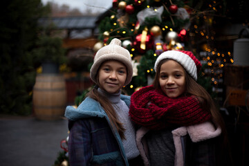 Two pretty pteteen girls  holds the gift  outdoor at the background decorated christmas tree with lights garland on New Year's Eve holidays. Kid dressed warm jacket, red knitted scarf , hat