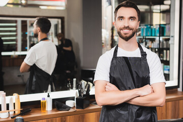 Cheerful hairdresser looking at camera while standing with crossed arms on blurred background