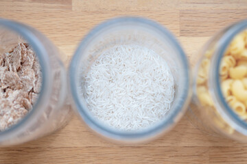 A glass jar with a long grainy white basmati and Jasmine rice in the center of the frame against a light wooden table top close-up view. Storing bulk food in the kitchen at home