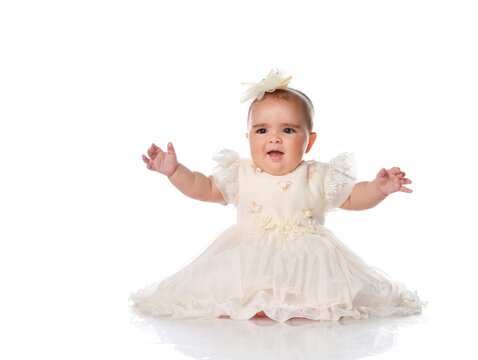 beautiful baby child in a festive dress and a headband, sitting on the floor and joyfully raised her hands. shot on a white background.