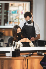 mirror reflection of hairdresser in face shield near woman in medical mask with hairdressing tools on blurred foreground