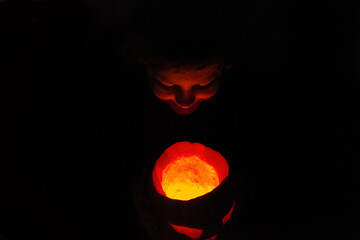 the face of a young boy looks into an open Halloween Jack-o'-lantern carved pumpkin glowing with...