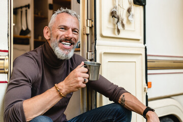 Smiling happy senior male traveler with beard and tattoo drinking coffee in the doorway of his caravan