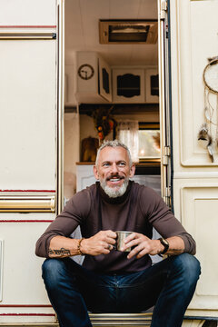 Vertical Shot Of A Mature Nice-looking Man Sitting On The Doorway Of His Mobile Home Smiling And Holding A Mug Of Tea: Front View