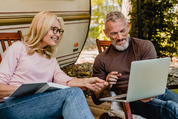 Mature handsome grey-haired man showing his blonde wife something amusing on his laptop in their garden
