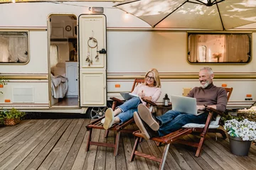 Foto op Plexiglas Pleased happy aged couple reading a book and using laptop while relaxing on the deck chairs near the camper van © InsideCreativeHouse