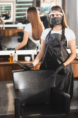 Fototapeta na wymiar hairdresser in apron and face shield holding scissors and comb while pointing with finger at armchair on blurred background
