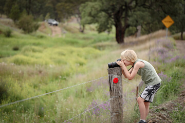 Little boy outdoors in pretty country location leaning on fence taking photos with dslr full frame camera