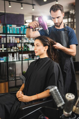 young hairdresser drying hair of woman near combs on blurred foreground