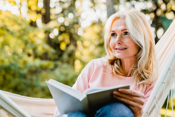 Beautiful senior blonde woman reading book and sitting in hammock in the garden - Powered by Adobe