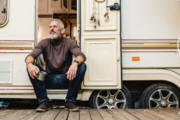 Portrait of handsome senior man with a beard sitting on the doorstep of the motor home holding iron...