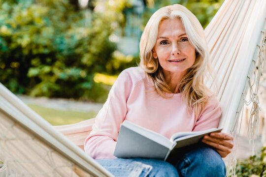 Nice-looking mature blonde woman resting in hammock in the garden with a book