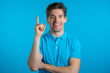 Smiling happy guy showing eureka gesture. Portrait of young man having idea moment pointing finger up on blue studio background. 