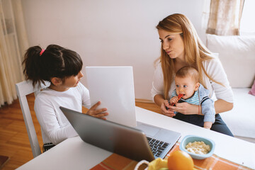 Mother working on laptop, holding baby boy and daughter learning 