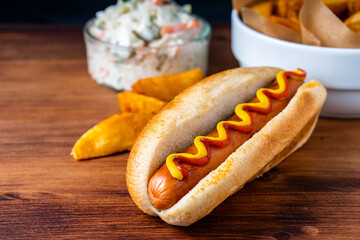 American traditional fast food Hotdog with fresh organic bread bun white wheat and black charcoal sausage ketchup mustard for meal with roasted onion pickle and French fries wedges and coleslaw