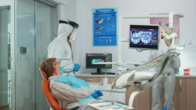 Dentist in coverall showing patient situation of teeth x-ray image during global pandemic. Assistant and doctor talking with senior woman wearing protection suit, face shield, mask and gloves.