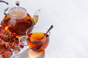 Sunny teatime with rosehip tea in transparent teapot with cup, autumn branch in bright cozy sun beams white wood table top view, copy space.