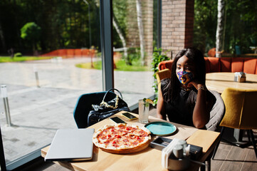 Fashionable african american woman, wear hand made face mask sitting at restaurant while eat pizza. New normal life.