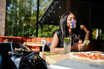 Fashionable african american woman, wear hand made face mask sitting at restaurant, look at mobile...
