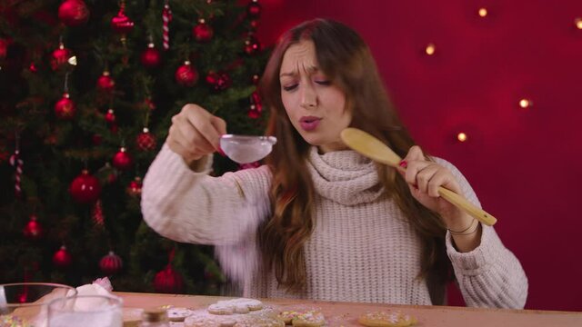 Cute long-haired girl sings song using wooden spatula instead of microphone on Xmas decoration with Christmas tree decorating handmade gingerbread. self-gifter Christmas bakery, festive mood concept.