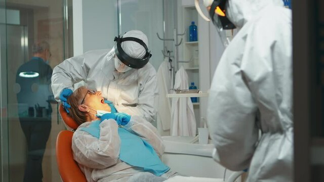 Dentist with face shiled treating teeth to senior woman patient in dental clinic during covid-19 . Doctor and nurse working wearing coverall, protection suit, mask, gloves in stomatological office.