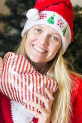 Christmas holiday portrait of cheerful young woman holding a gift by the tree wearing red hat.