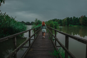 Back view of a female walking on a wooden bridge through green bushy lake with a lot of leaves on a murky scary cloudy day.