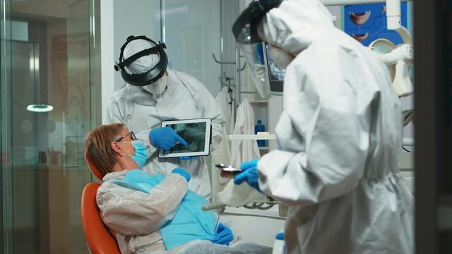 Stomatologist in protective suit reviewing x-ray of tooth with senior patient explaining treatment using tablet in covisd-19 pandemic. Medical team wearing face shield, coverall, mask and gloves.