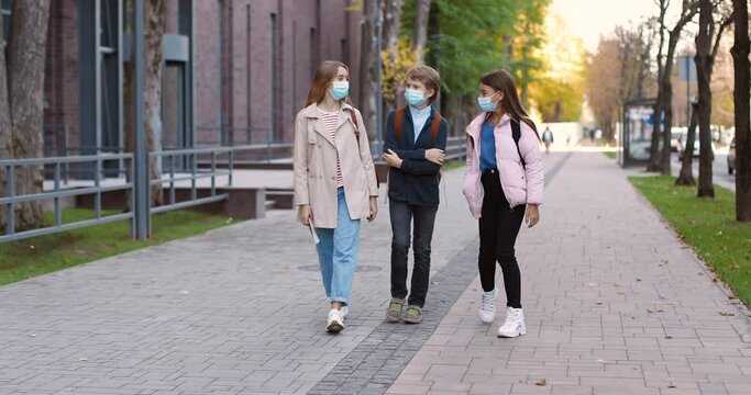 Caucasian teen schoolchildren walking after lessons and chatting in good mood. Boy and beautiful happy girls in masks talking. Joyful college students and friends on street. School concept