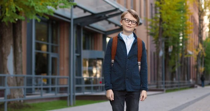Portrait of joyful Caucasian boy pupil in glasses in good mood standing outdoor near school. Happy male student with backpack smiling to camera on street in town. Education concept