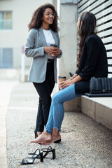 Two beautiful businesswomen talking while drinking coffee sitting on a bench on the street.