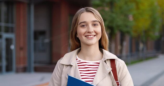 Close up of joyful Caucasian girl standing outdoor and smiling to camera. Portrait of cheerful female pupil with smile on face on street near school. Positive emotions. School concept